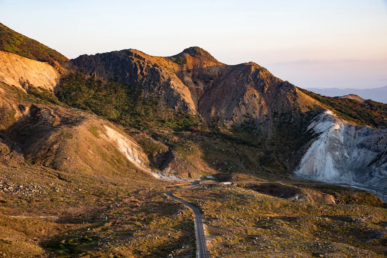 夕方の光を受ける火山の山肌と、斜面を縫って上る磐梯吾妻スカイライン